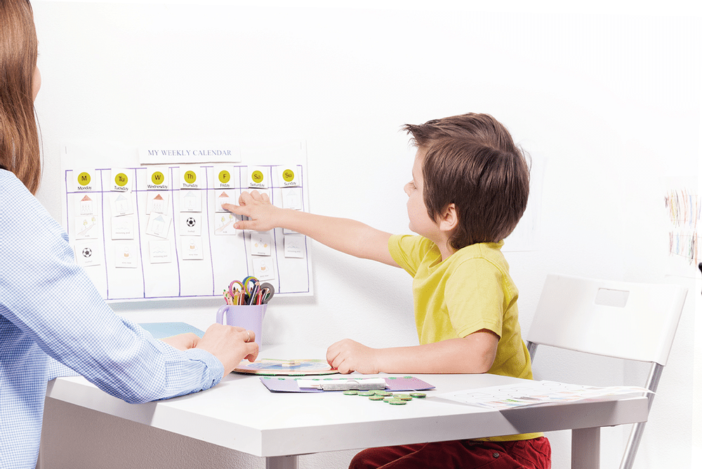 Young boy wearing a yellow shirt points with his right arm to a white board as he attempts to complete math problems.