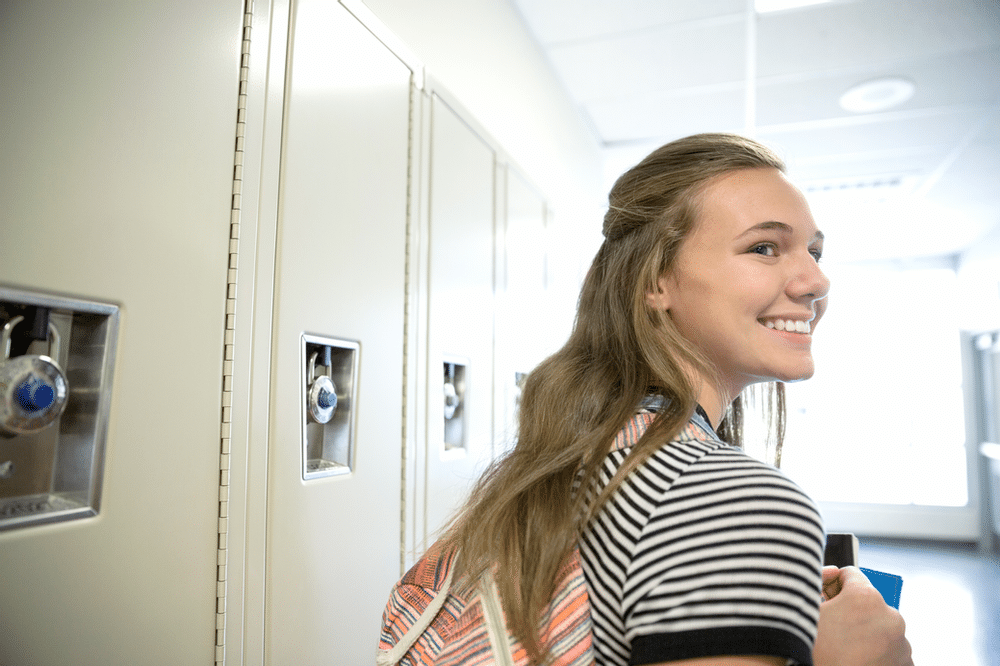 High-school aged, female-presenting white student smiles in front of lockers.