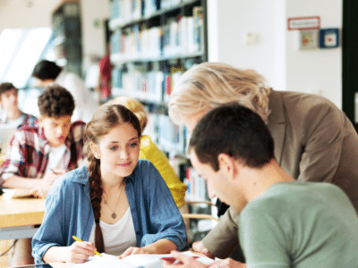 Two high school-aged students, one female-presenting, the other male-presenting, sit at a table with open books. An older female-presenting figure with face obscured leans between them.