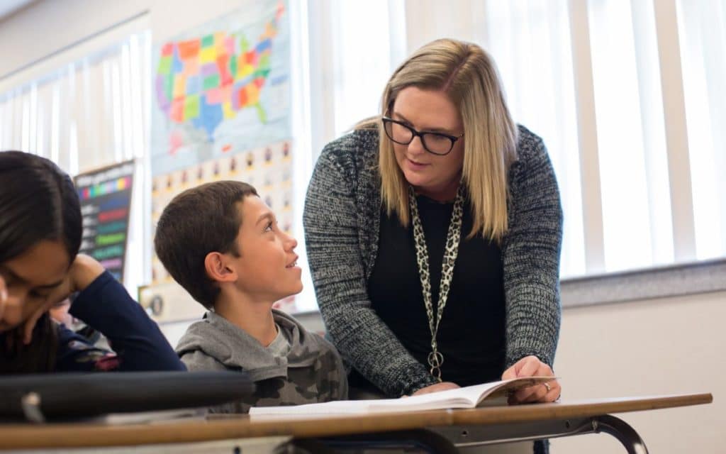 2 students seated in a classroom, conversing with their teacher over a book