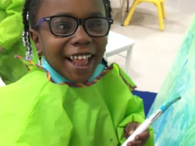 Kindergarden student with visual impairments smiles at the camera while holding a paintbrush. Her artwork of blue and green paint is on a canvas to the left of her.
