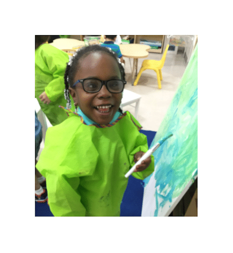 Kindergarden student with visual impairments smiles at the camera while holding a paintbrush. Her artwork of blue and green paint is on a canvas to the left of her.