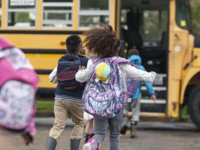 A multi-ethnic group of elementary age children are getting on a school bus. The kids' backs are to the camera. They are running towards the school bus which is parked with its door open. It's a rainy day and the kids are wearing jackets, rain boots and backpacks.