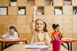 Young student with pigtails raising her hand at her desk in a classroom.