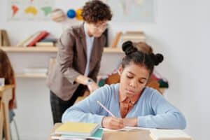 Student writing with a pencil at a desk with teacher in the background